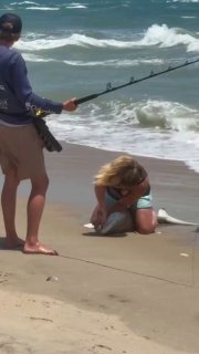 Pliers in hand, a woman kneels next to a shark on the beach and pulls a fishing lure from its jaws before sending it back into the water.